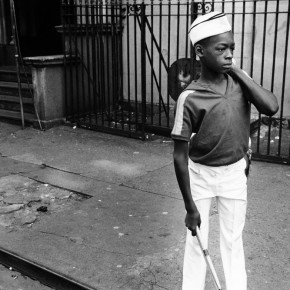 boy in marching band outfit standing on sidewalk in Harlem, carrying drum over his shoulder and holding a drum stick