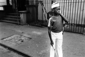 boy in marching band outfit standing on sidewalk in Harlem, carrying drum over his shoulder and holding a drum stick