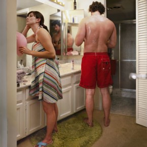 woman wrapped in towel looking in mirror; man in swim shorts stands next to her with his back turned