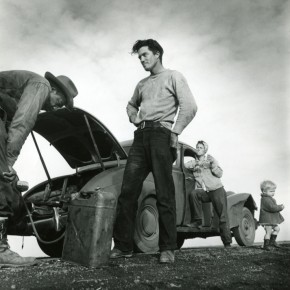 man standing next to car; man in hat peers into open trunk; woman and child stand behind them