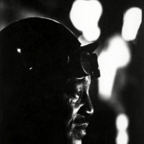 profile of man wearing a hardhat and goggles on his forehead in the dark with bright lights in background