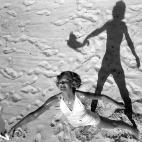 semi-aerial view of woman standing on beach, holding out a handkerchief with her shadow behind her