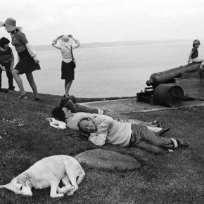 people sleeping, sitting and standing on grass next to body of water; child sits on cannon