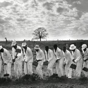 row of men in a field wearing matching outfits, holding shovels and digging